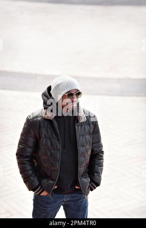 Street fashion, uomo elegante ritratto in giacca nera in pelle, felpa con cappuccio, cappello lavorato a maglia e occhiali da sole in piedi, mani in tasca Foto Stock