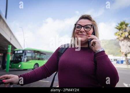 Una giovane donna sorridente fa una telefonata mentre aspetta l'autobus alla stazione Foto Stock