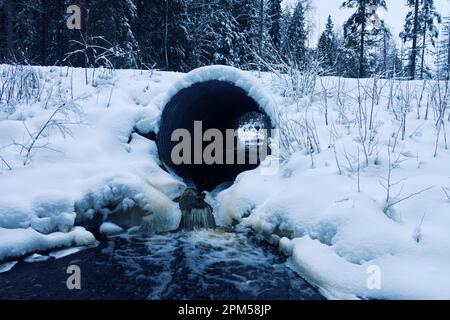 Il canale sotto la strada forestale è coperto di neve in inverno, ma lo scarico d'acqua continua, ghiaccio - ruscello libero Foto Stock