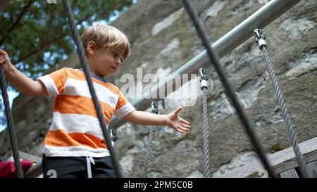 Bambino attivo che soggiorna in equilibrio attraversando il ponte del parco giochi all'esterno. Un ragazzino che tiene i cavi metallici attraversa la struttura. Capretto extercising outdoo Foto Stock