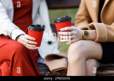 due donne che tengono tazze di carta con bevande calde seduto su una panca Foto Stock