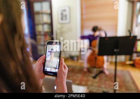 Un bambino con il cellulare registra il fratello che suona il violoncello a casa Foto Stock