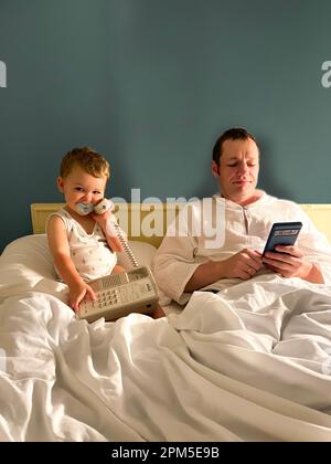 Padre e figlio con succhietto tengono il telefono a letto Foto Stock