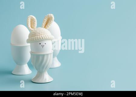 uovo di pasqua in cappello all'uncinetto con le orecchie da coniglietto nel portuovo su blu Foto Stock