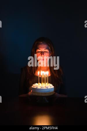 Donna attraente che soffia le candele sulla torta di compleanno in camera scura. Foto Stock