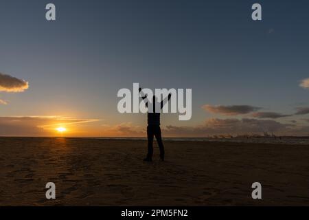 Giovane uomo, in piedi, alzando le braccia, guardando il tramonto sulla spiaggia Foto Stock