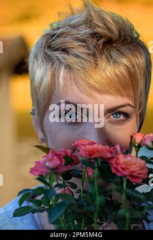 Ritratto vividamente colorato di una giovane donna con corti capelli biondi all'aperto in natura, odorando e apprezzando piccoli fiori rosa. Un regalo da sua madre Foto Stock