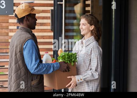 Vista laterale ritratto di consegna sorridente uomo scatola di consegna con alimentari freschi a giovane donna all'aperto al portico casa Foto Stock
