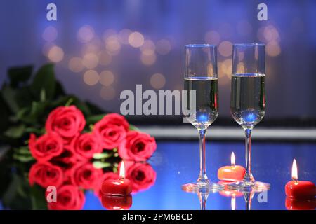 Bicchieri di champagne, candele e rose sul tavolo contro luci sfocate. Cena romantica per San Valentino Foto Stock