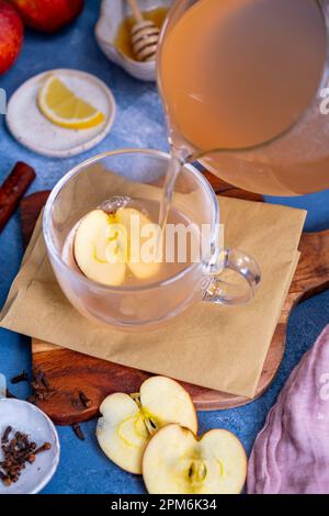 Il tè di mele viene versato in una tazza di vetro, le fette di mela, i chiodi di garofano, le fette di limone, il miele intorno esso. Foto Stock