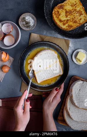Una fetta di pane bianco viene immersa nel composto di uova e latte in una ciotola e una fetta di toast alla francese su un piatto sul lato. Foto Stock