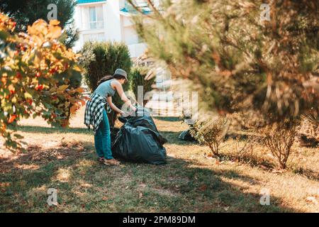 Il concetto di inquinamento ambientale e giorno della Terra. Donna volontaria che fa la pulizia nel Parco di fronte alla casa. Sta mettendo i sacchetti della spazzatura Foto Stock