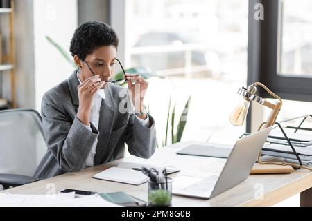 elegante afroamericana businesswoman mettere su occhiali mentre si siede vicino a notebook e notebook vuoto in ufficio, immagine stock Foto Stock