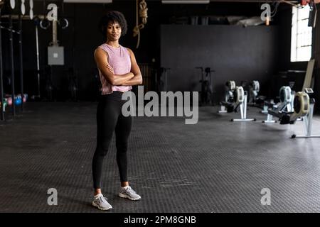 Ritratto completo di giovane donna fidata biraciale con braccia incrociate in piedi in palestra Foto Stock