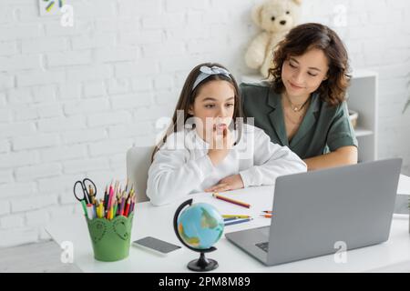 Donna positiva che guarda la bambina pre parlare durante la lezione di terapia vocale sul laptop a casa, immagine stock Foto Stock