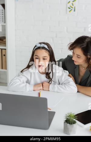 mamma sorridente che guarda la figlia che parla durante una lezione di terapia vocale sul computer portatile a casa, immagine di scorta Foto Stock