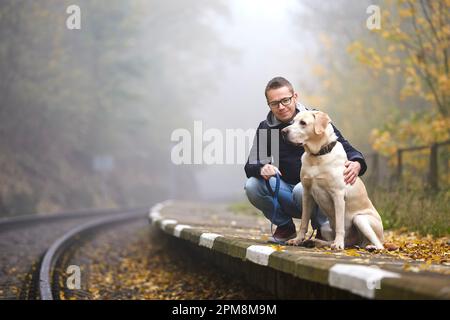 L'uomo con il cane al guinzaglio sta aspettando sulla piattaforma della stazione ferroviaria. Il proprietario dell'animale domestico e il suo labrador Retriever insieme durante il viaggio in treno. Foto Stock