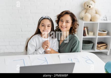 Donna e figlia allegra guardando la macchina fotografica vicino a carte con lettere sulla scrivania, immagine di scorta Foto Stock