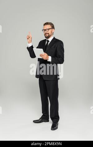 intera lunghezza di uomo d'affari in abbigliamento formale e occhiali che tengono il tablet digitale mentre puntano con il dito sul grigio, immagine stock Foto Stock