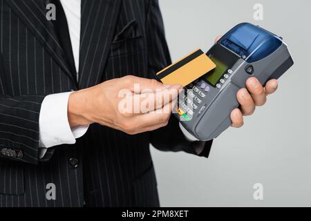vista ritagliata di un uomo d'affari in tuta con carta di credito vicino al terminale di pagamento isolato su grigio, immagine stock Foto Stock