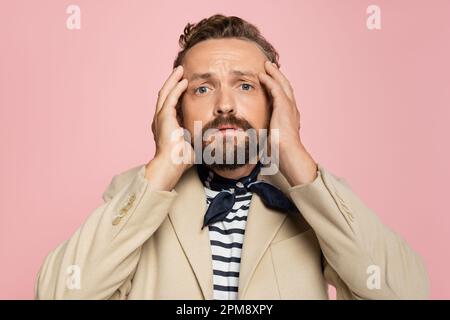 uomo stressato in blazer e sciarpa collo guardando la fotocamera mentre tenendo la testa isolata su rosa, immagine stock Foto Stock