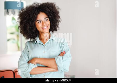 Donna sorridente ricciola in piedi in appartamento moderno e guardando da parte sognante. Giovane donna positiva con capelli afro, orgogliosa e di successo Foto Stock