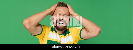 appassionato di sport emotivo in sciarpa a righe e t-shirt gialla tirando i capelli isolati su verde, banner, immagine stock Foto Stock