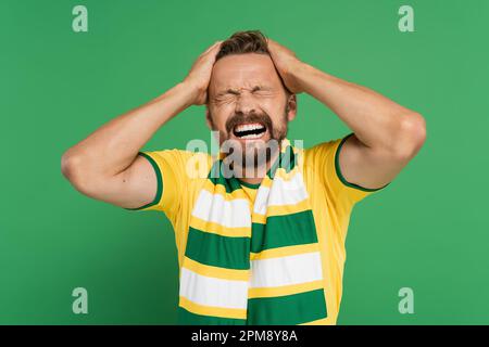 appassionato di sport emozionale in sciarpa a righe e t-shirt gialla che tira i capelli isolati su verde, immagine stock Foto Stock