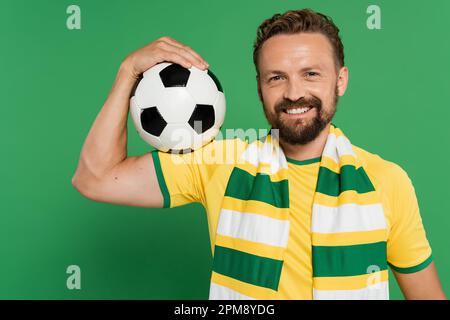 sorridente tifoso di calcio in sciarpa a righe e t-shirt gialla che tiene il calcio isolato su verde, immagine stock Foto Stock