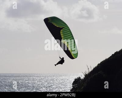 Un parapendio vola su una scena oceanica con un affioramento di terra alla sua destra.Backlit silhouette.Sport.Recreation Foto Stock