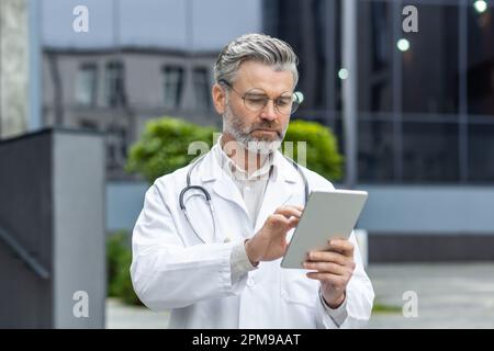 Medicina online. Un vecchio medico maschio dai capelli grigi si trova fuori dalla clinica in un cappotto bianco e usa una compressa. Chiamate, chiamate, SMS, conversazioni. Foto Stock