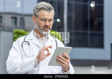 Medicina online. Un medico di sesso maschile senior si trova fuori dalla clinica in un cappotto bianco e tiene una tavoletta nelle mani. Parla con i pazienti, consulta una videochiamata, punta la telecamera con le dita. Foto Stock