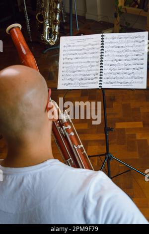 Immagine verticale vista posteriore dell'uomo calvo vestito di bianco, seduto sul divano nel soggiorno, studiando musica classica e suonando fagotto, leggendo il mus del foglio Foto Stock