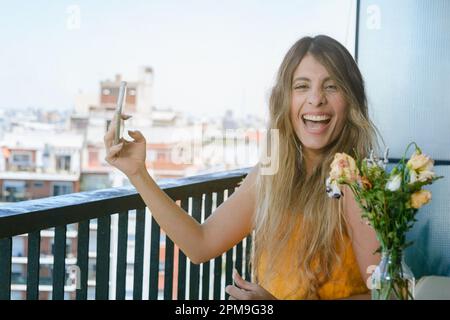 giovane donna latina di etnia colombiana seduta sul balcone del suo appartamento tenendo il telefono e guardando la fotocamera sorridendo molto felice, techn Foto Stock