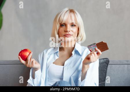 Giovane ragazza bionda premurosa sceglie tra cioccolato e mela. Seduta sul divano. Foto Stock