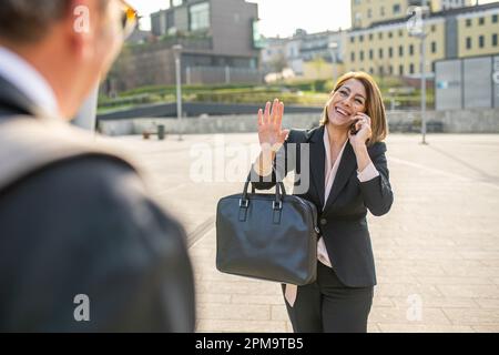 Una donna matura che utilizza lo smartphone durante una chiamata incontra la sua collega di fronte all'ufficio, con la luce del sole sul retro Foto Stock