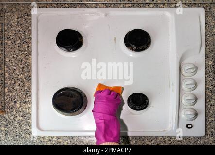 Una mano in un guanto in lattice rosa con una spugna con schiuma detergente lava la stufa a gas in cucina. Foto Stock