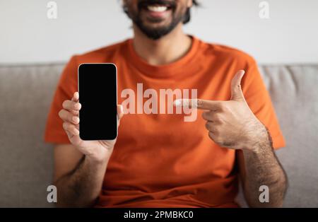 Giovane ragazzo medio orientale felice sul divano punta il dito contro lo smartphone con schermo vuoto nel salotto interno Foto Stock