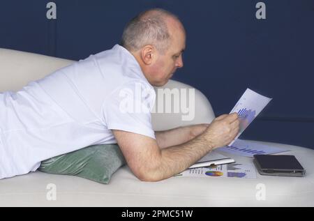 Un uomo d'affari si trova su un divano e guarda le tendenze dei dati su grafici e grafici. Lavoro a distanza Foto Stock