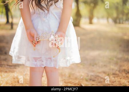 La bambina in vestito bianco è sulla schiena che tiene una corona principessa, in una foresta da sogno, spazio copia, tema giorno dei bambini. Foto Stock