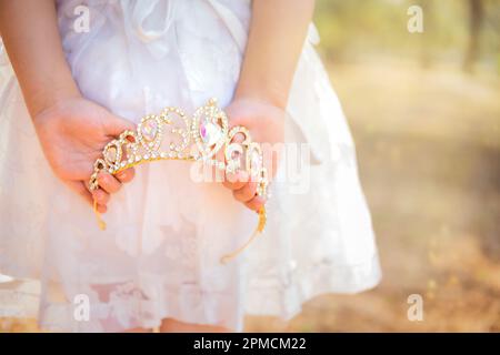 La bambina in vestito bianco è sulla schiena che tiene una corona principessa, in una foresta da sogno, spazio copia, tema giorno dei bambini. Foto Stock