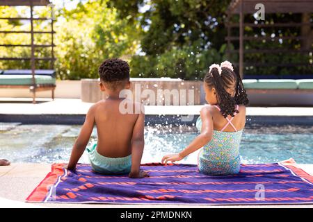 Vista posteriore dei fratelli afroamericani che spruzzano acqua in piscina mentre si siedono su un asciugamano a bordo piscina Foto Stock