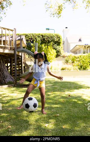 Intera lunghezza di ragazza afroamericana allegra che gioca il calcio sul campo erboso nel parco Foto Stock