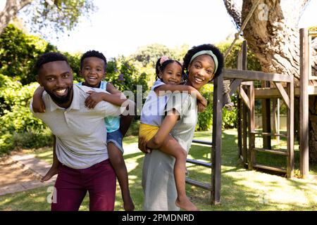 Felice afroamericano genitori piggybacking figlio e figlia mentre in piedi in parco, copia spazio Foto Stock