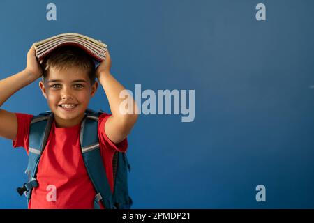 Ritratto di un ragazzo caucasico sorridente che porta libri in testa mentre si alza su sfondo blu Foto Stock