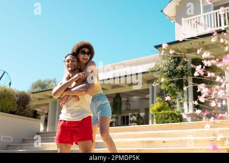 Felice coppia diversa in piedi fuori casa, abbracciando e sorridendo Foto Stock