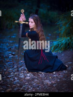 Giovane donna inginocchiata in fiaba Queen Costume Holding Broadsword | Moody Foto Stock