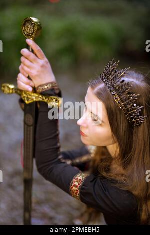 Giovane donna inginocchiata in fiaba Queen Costume Holding Broadsword | Moody Foto Stock