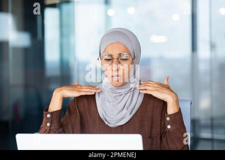 Calore e calore in ufficio, donna d'affari in hijab agitando le mani cercando di rinfrescarsi e raffreddarsi, aria condizionata rotto sul posto di lavoro. Foto Stock