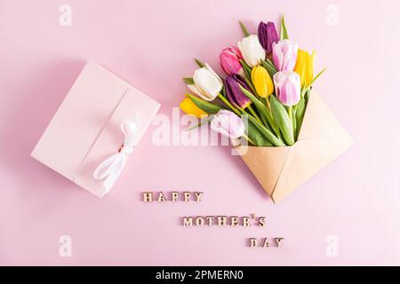 Una composizione festiva per la Festa della mamma. Bellissimi tulipani primavera colorati in una busta artigianale e una scatola rosa con un regalo. vista dall'alto. testo Foto Stock